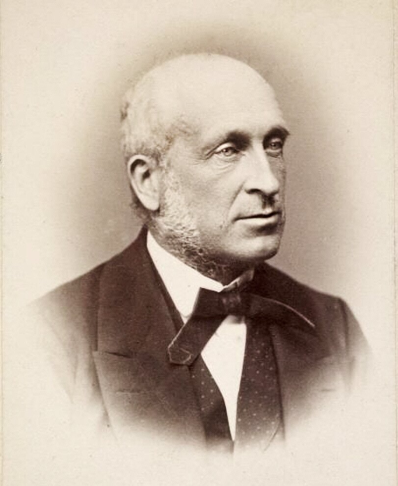 Zdjęcie przedstawia portret starszego mężczyzny. Ukazany jest jego prawy półprofil. Mężczyzna jest łysy, ma zmarszczki na twarzy oraz siwe bokobrody. Ubrany jest w garnitur z przewiązaną muchą.