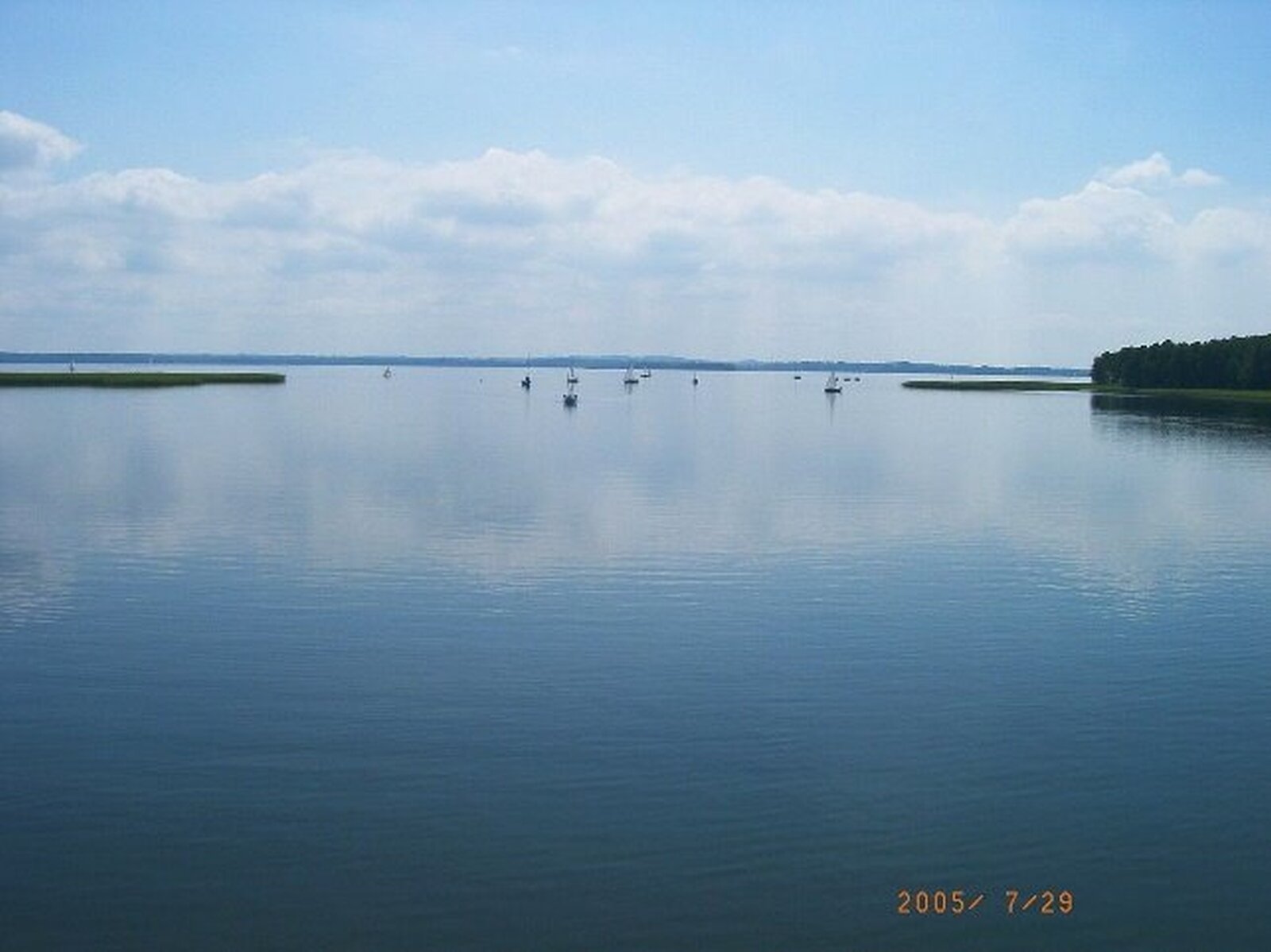 Zdjęcie przedstawia rozległe jezioro. Gładka tafla wody, pośrodku kilka pływających żaglówek. W tle i po prawej stronie linia otaczającego jezioro lasu.