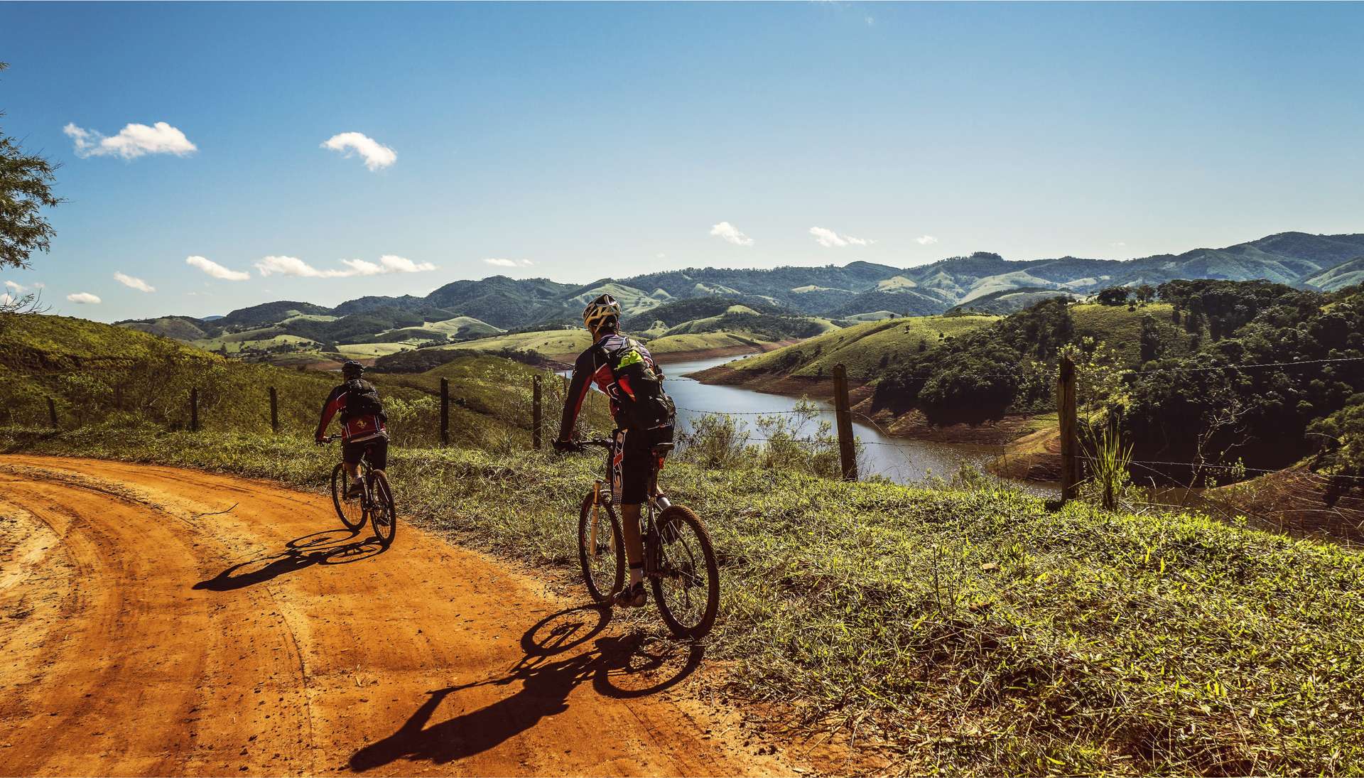 Zdjęcie przedstawia na pierwszym planie dwóch mężczyzn na rowerach górskich. Ubrani są w kolarskie stroje, na głowach mają kaski ochronne. Mężczyźni jadą piaskową drogą. Na drugim planie widoczny jest krajobraz. Widoczna jest rzeka, góry oraz prawie bezchmurne niebo.