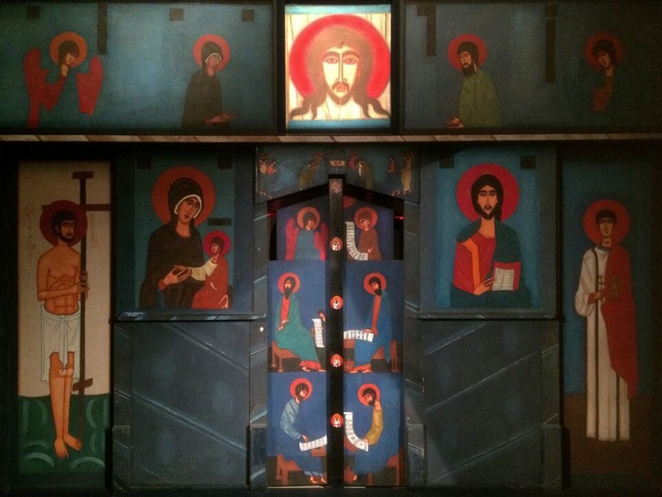 Ilustracja przedstawia malarstwo naścienne tzw. Ikonostas Jerzego Nowosielskiego pt. „Szafirowy Ikonostas”. Na powierzchni znajdują się postacie świętych. Cały górny rząd składa się z pięciu postaci - po dwie postaciach z każdej strony, a w centrum widoczna jest głowa Chrystusa. Rząd niżej widoczna jest scena - postać półnagiego świętego, ubranego w białą szatę, stojącego z głównym krzyżem, Maryję z dzieciątkiem. Kobieta ubrana jest w ciemną szatę, dzieciątko w biało‑czerwoną szatę. Dorosłego Chrystusa siedzącego i trzymającego kartkę. Chrystus ubrany jest w zielono‑czerwoną szatę, a także ma brodę na twarzy. Obok Chrystusa po prawej stronie widoczny jest kolejny święty ubrany w biało‑czerwoną szatę. Pośrodku malowidła ściennego ukazany jest przedmiot przypominający drzwi na których widoczne są postacie świętych.