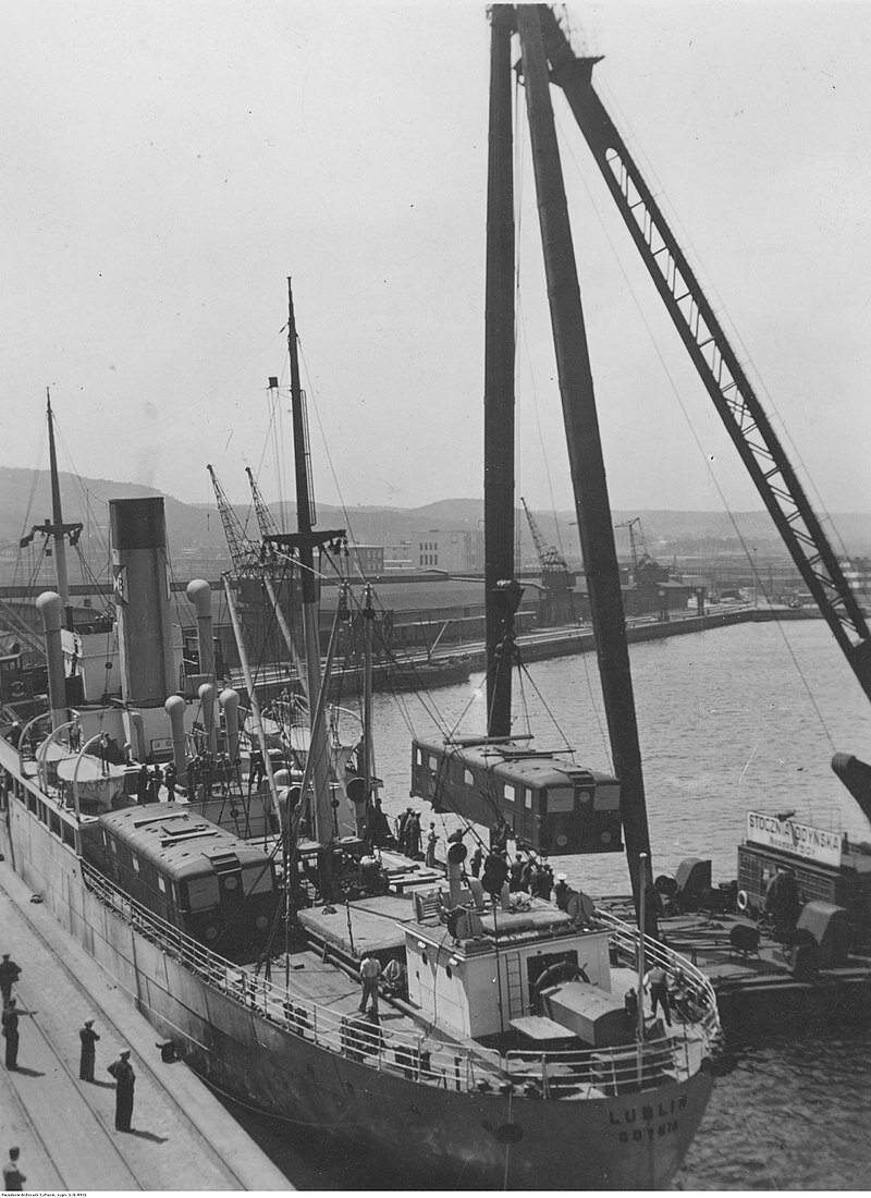 Zdjęcie przedstawia statek podczas załadunku. Dźwig przenosi transportowany pojazd na pokład statku. Przygląda się temu grupa osób, między innymi załoga statku. Na podstawie dźwigu napis „STOCZNIA GDYŃSKA”. Drugi taki sam pojazd już został dostarczony na pokład. Na dalszej części pokładu widać szalupy ratunkowe. W tle widoczne są budynki stoczniowe. Ilustracja jest czarno‑biała.