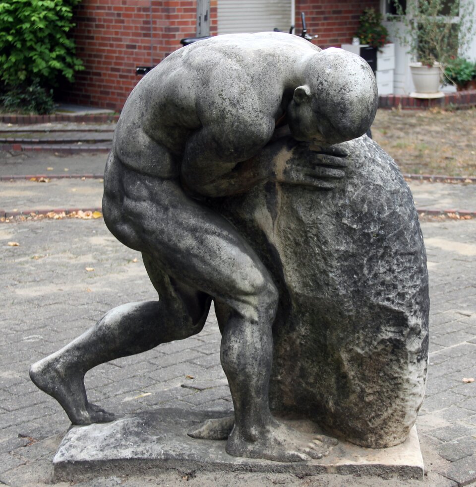 Fotografia przedstawia rzeźbę nagiego, muskularnego mężczyzny. Jest to osoba w sile wieku. Pochyla się nad dużym głazem w kształcie jaja. Postać napina mięśnie, usiłując podnieść kamień.