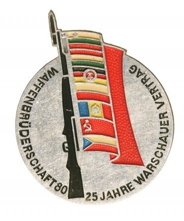 Ilustracja przedstawia okrągły znaczek, na którym znajduje się pionowo ustawiony karabin z ośmioma flagami zawieszonymi na lufie. Na krawędzi napis w języku niemieckim.