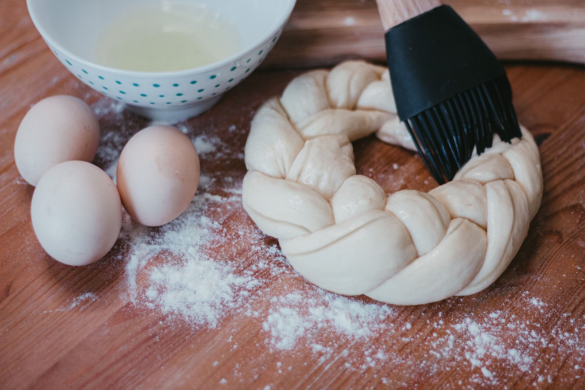 Zdjęcie przedstawia krok ósmy formowania wianka debrzeńskiego. Ujęcie wykonane jest od góry pod skosem. Widoczny jest ciemnobrązowy blat na którym w środkowej części grafiki znajduje się owalny zapleciony wianek z ciasta. Dookoła wianka rozsypana jest biała mąka. Wyrób smarowany jest przezroczystym jajkiem za pomocą silikonowego czarnego pędzelka kuchennego. W lewej części zdjęcia widoczne są trzy jajka leżące na blacie. Nad nimi znajduje się biała miseczka w małe niebieskie kropki w środku której dostrzec można białka jajek. W górnej prawej części zdjęcia znajduje się fragment drewnianej jasnobrązowej deski.