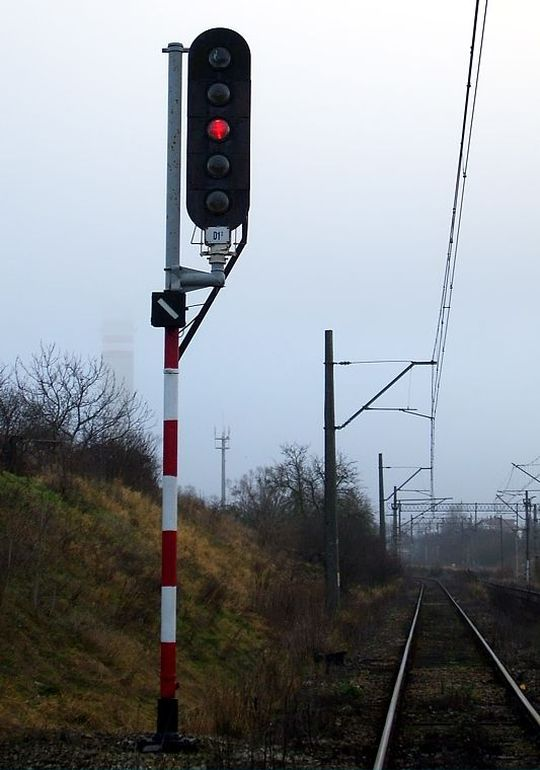 Zdjęcie przedstawia semafor będący elementem semaforowych obwodów świateł sygnałowych.  Stoi on obok torów i  posiada pięć świateł umieszczonych pionowo, a sam semafor przymocowany jest do masztu pomalowanego w biało czerwone pasy. Na semaforze środkowe światło świeci się na czerwono. 