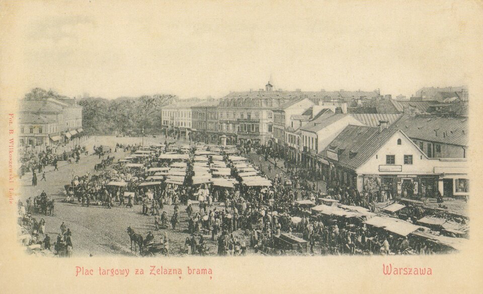Obraz przedstawia plac targowy za Żelazną Bramą w Warszawie. Na pierwszym planie widoczne są rzędy stoisk z zadaszeniem oraz tłum ludzi biorących udział w handlu. W tle znajdują się budynki mieszkalne i usługowe z dwuspadowymi dachami. Całość utrzymana jest w tonacji sepii.