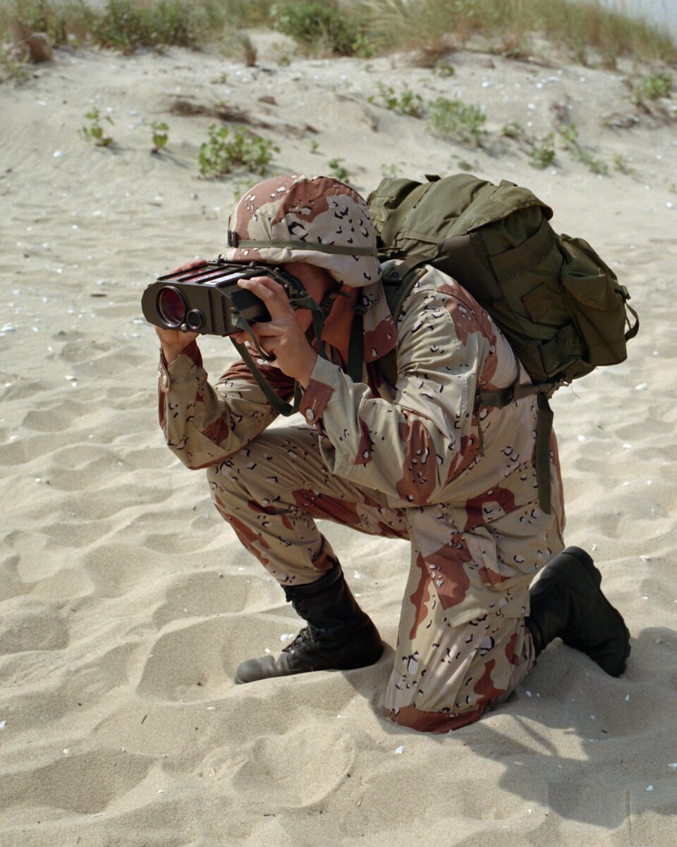 Kolorowa fotografia przedstawia klęczącego na jednym kolanie żołnierza na plaży. Jest ubrany w maskujący mundur w beżowo‑brązowo‑czarne plamy. Na plecach nosi ciemnozielony plecak. Na głowie ma hełm, na nogach wysokie buty, patrzy przez lornetkę.