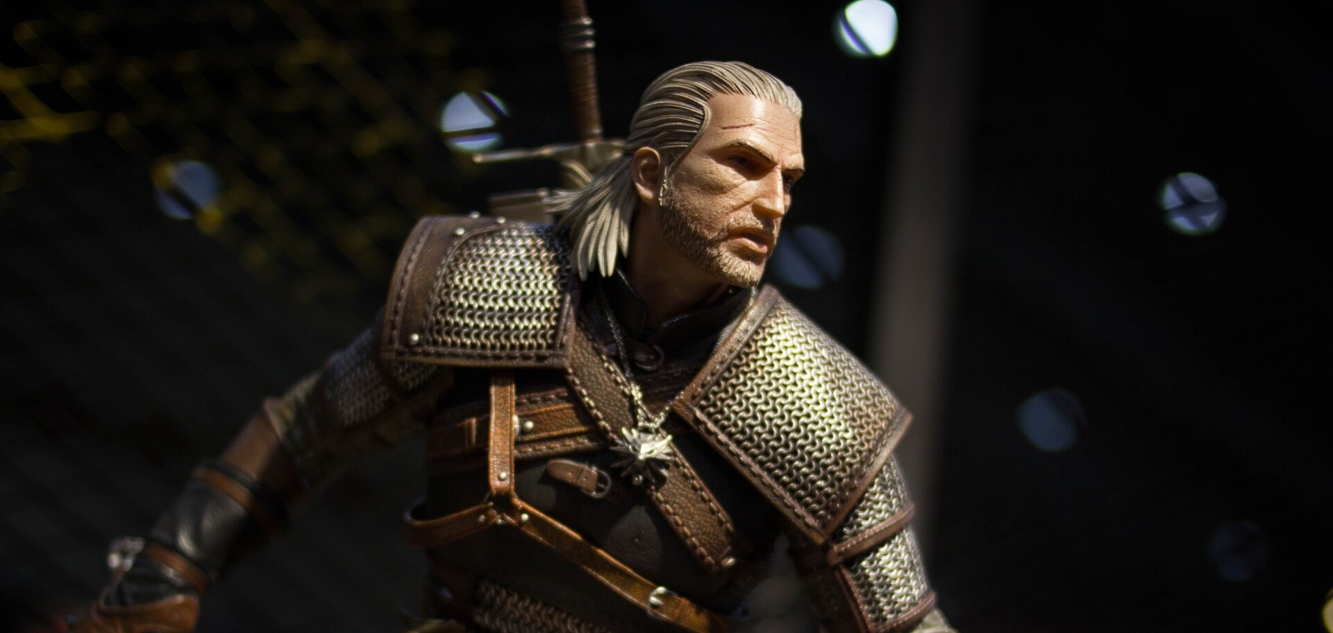 Zdjęcie przedstawia figurkę Geralta z Rivii. To dojrzały mężczyzna w rycerskiej zbroi, z mieczem na plecach. Mężczyzna ma białe włosy związane w kucyk, sięgające ramion. Na czole ma bliznę.
