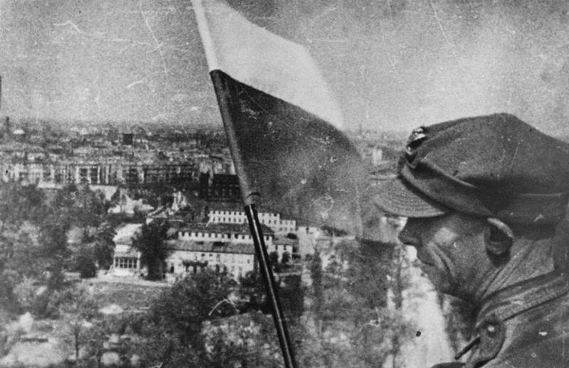 Zdjęcie przedstawia polskiego żołnierza wieszającego flagę polską na szczycie Bramy Brandenburskiej w Berlinie. W tle widać panoramę Berlina.