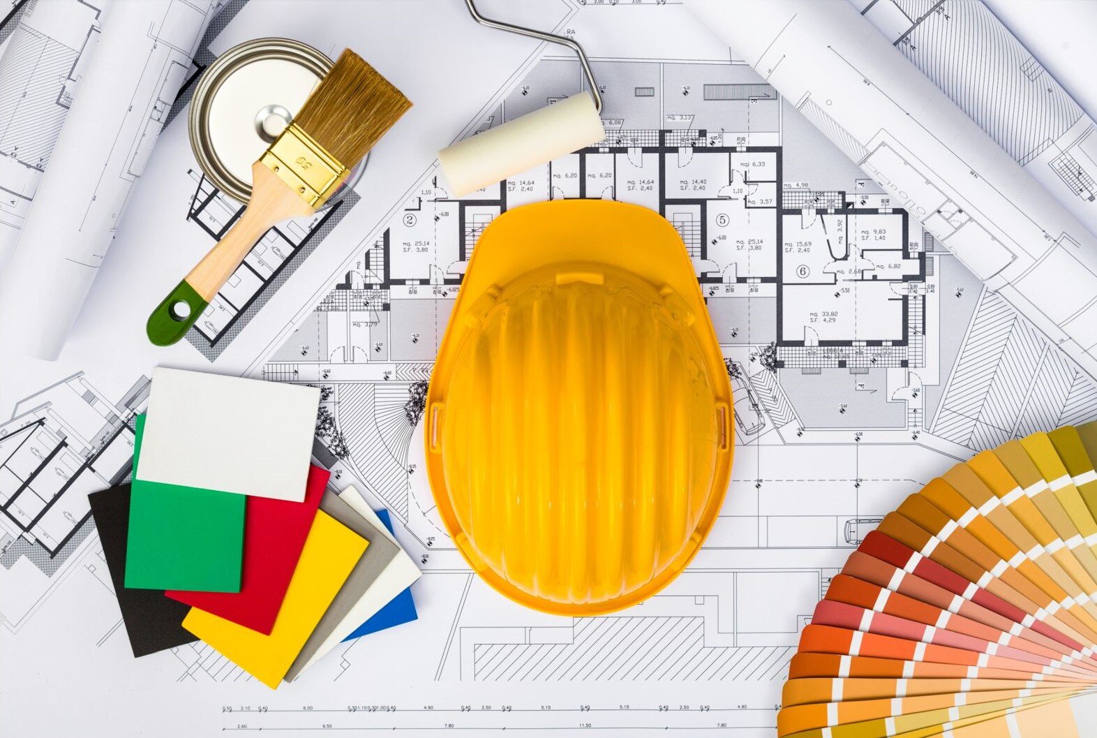Zdjęcie ukazuje stół kreślarski z planami mieszkania, domu. Na nim leżą kolorowe kartoniki, szeroki pędzel na puszce z farbą i kask ochronny stosowny na budowie. W rogu rozłożone zostały w formie wachlarza wzorniki kolorów do malowania ścian.