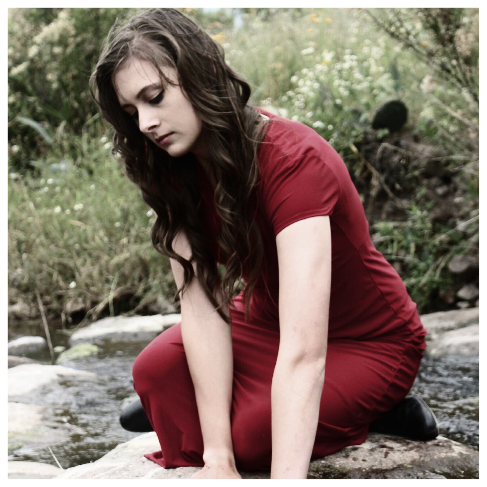 Zdjęcie przedstawia młodą kobietę siedzącą na skale nad rzeką. Rękę moczy w wodzie. Kobieta ma długie, brązowe, falowane włosy. Jest szczupła.