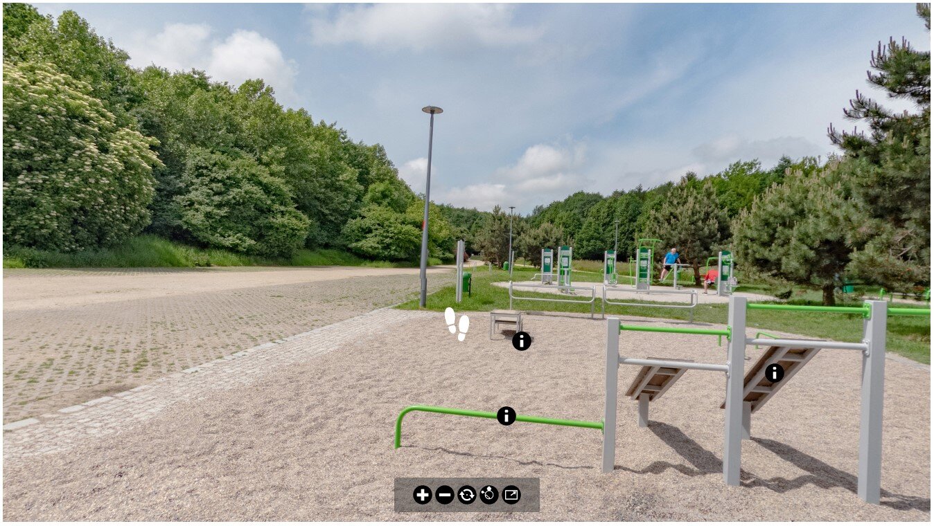 Ilustracja przedstawia przykładowy widok panoramy wycieczki wirtualnej. Przedstawia ona teren, na którym znajduje się siłownia terenowa. Na środku ekranu umieszczony został piktogram stóp. Jego wciśnięcie spowoduje przejście do kolejnej panoramy, z której składa się dana wycieczka. 