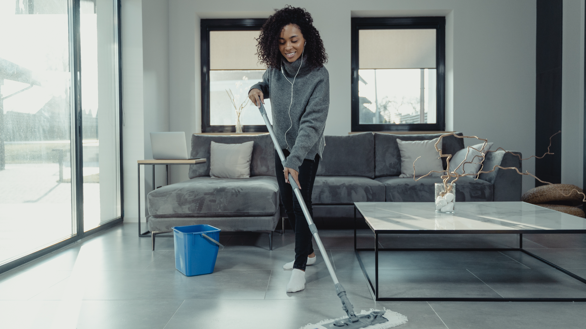 Zdjęcie przedstawia uśmiechniętą, młodą kobietę o ciemnej karnacji z bujnymi kręconymi włosami. Kobieta zmywa mopem podłogę w salonie.