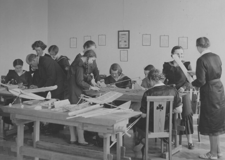 Fotografia przedstawia jedną z klas podczas lekcji techniki. Klasa składa się z dziewczyn, które są w trakcie konstruowania modeli samolotów. Przy jednej z nich stoi nauczyciel i pomaga w pracy. Na ścianach wiszą obrazki. Uczennice rozmawiają ze sobą i uśmiechają się.