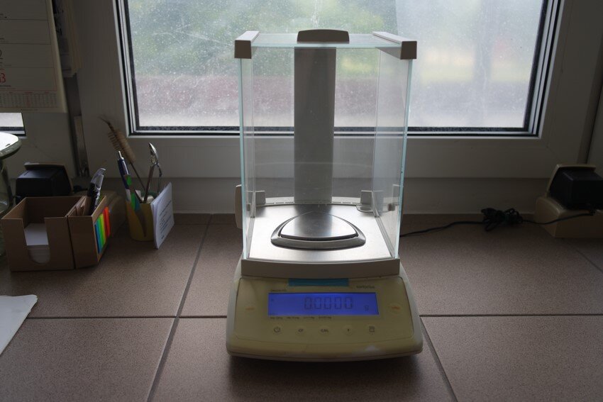 Zdjęcie przedstawia wagę laboratoryjną do oznaczania gramatury i naważek. Waga posiada podstawę z przyciskami sterującymi i wyświetlaczem oraz zabezpieczone szklanym kloszem miejsce do ważenia substancji. 