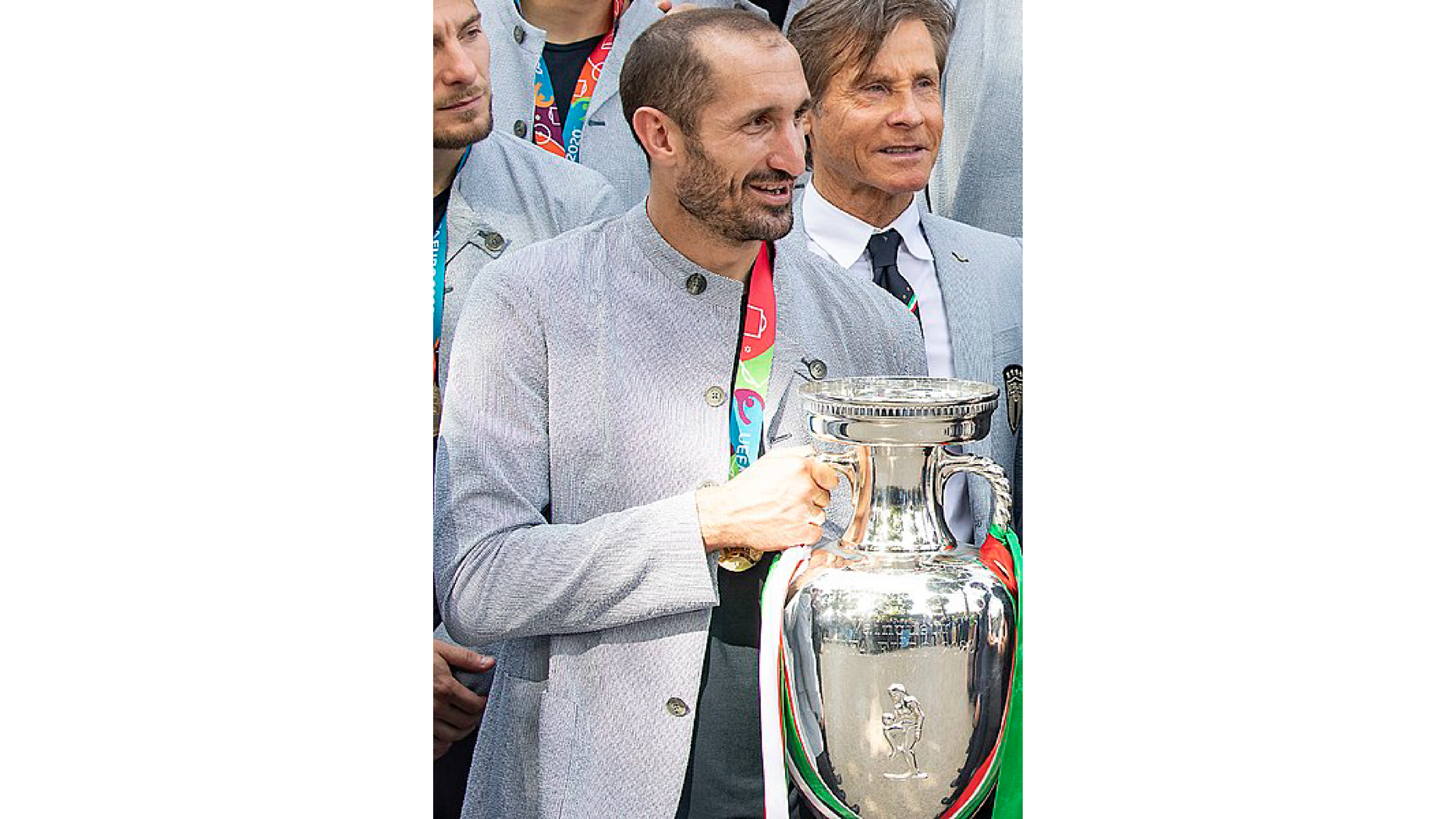 Zdjęcie przedstawia Giorgio Chielliniego, znanego włoskiego piłkarza trzymającego puchar. Piłkarz ubrany jest w marynarkę. Za piłkarzem stoją inni zawodnicy. 