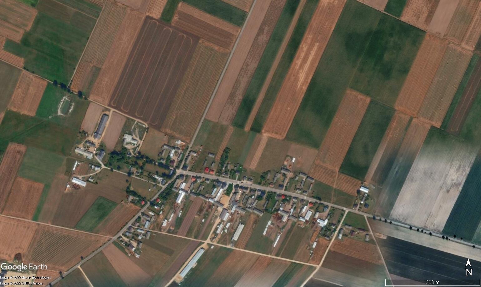 Ilustracja przedstawia gospodarstwa rolne w Polsce które skupione są w niewielkich miejscowościach, otoczonych polami uprawnymi. W jednej części obszaru, przy głównej drodze, zgromadzone są budynki mieszkalne i gospodarcze. Za nimi rozciągają się pola uprawne. Mają one różne długości, są jednak przeważnie w prostokątnych kształtach.