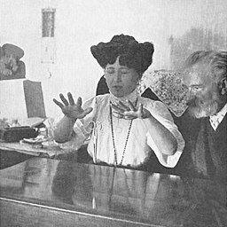 Zdjęcie przedstawia dwoje ludzi siedzących przy stole. To starsza kobieta z długimi, upiętymi na głowie włosami, która trzyma uniesione wysoko nad stołem ręce i starszy, brodaty mężczyzna w garniturze, który przypatruje się dłoniom kobiety.