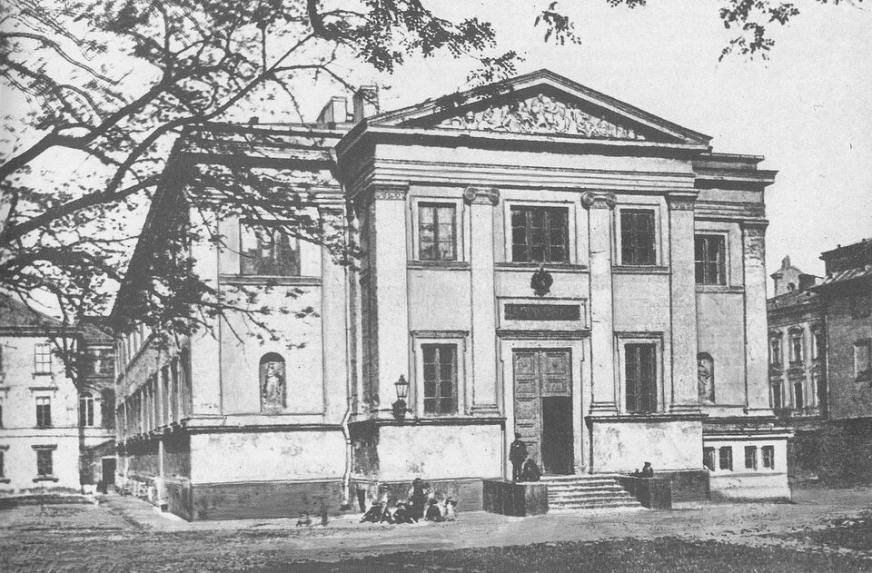 Ilustracja przedstawia budynek. Budynek jest dwukondygnacyjny. Do obiektu prowadzi kilka schodów. Na fasadzie są kolumny, a w dolnej części po lewej i prawej stronie nisze, w których stoją posągi. Fasadę zwieńcza fronton - trójkątny szczyt. Po prawej stronie budynku oraz z tyłu stoją inne budynki. 