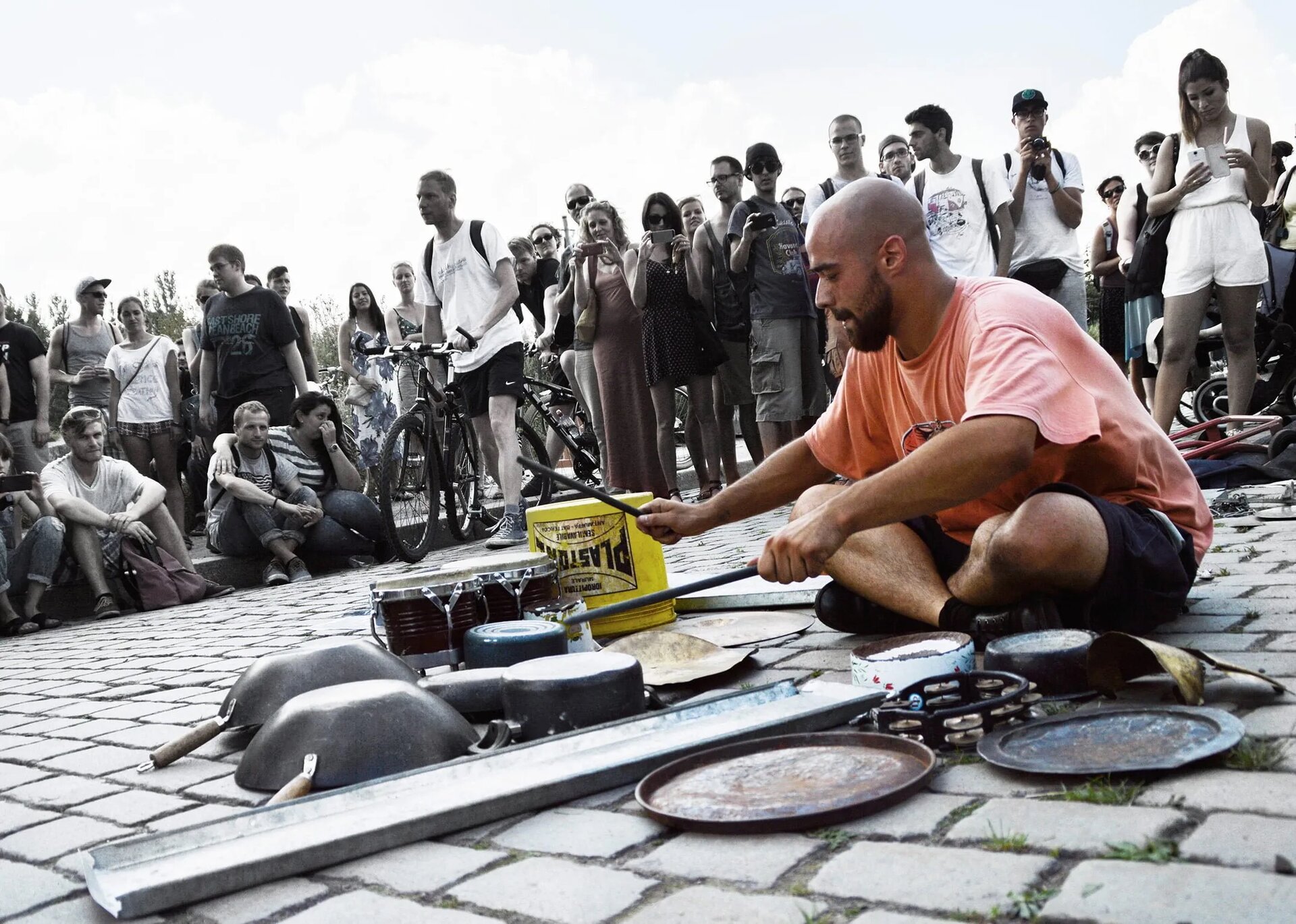 Na zdjęciu młody, łysy mężczyzna o ciemnej brodzie, siedzący na kostce brukowej po turecku, który został otoczony przez tłum gapiów. Mężczyzna który znalazł się w centrum uwagi jest artystą ulicznym i jest w trakcie odbywania występu. Gra pałeczkami na bębnie, ale część z jego instrumentów stanowi przedmioty kuchenne, na przykład garnki, talerze oraz woki. Część z obserwujących stoi, część siedzi na krawężniku, niektórzy robią zdjęcia.