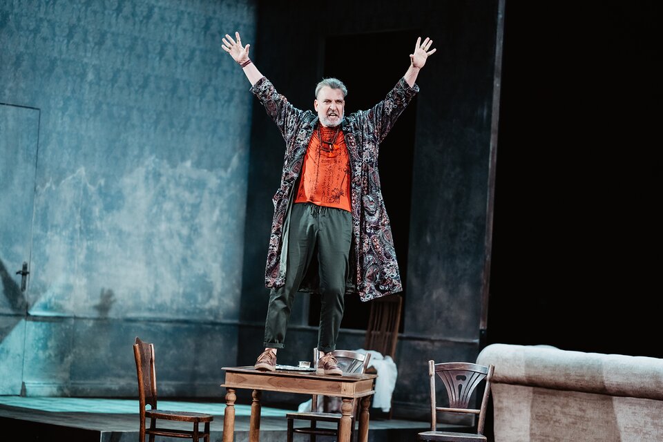 Na zdjęciu widać mężczyznę z podniesionymi do góry dłońmi stojącego na stole. Prawdopodobnie przemawia. Ubrany jest w brązowe sportowe buty, zielone spodnie, czerwoną wyszywaną koszulę, na którą opadają korale oraz kolorowy rozpięty szlafrok. W tle widać niebieską ścianę. 