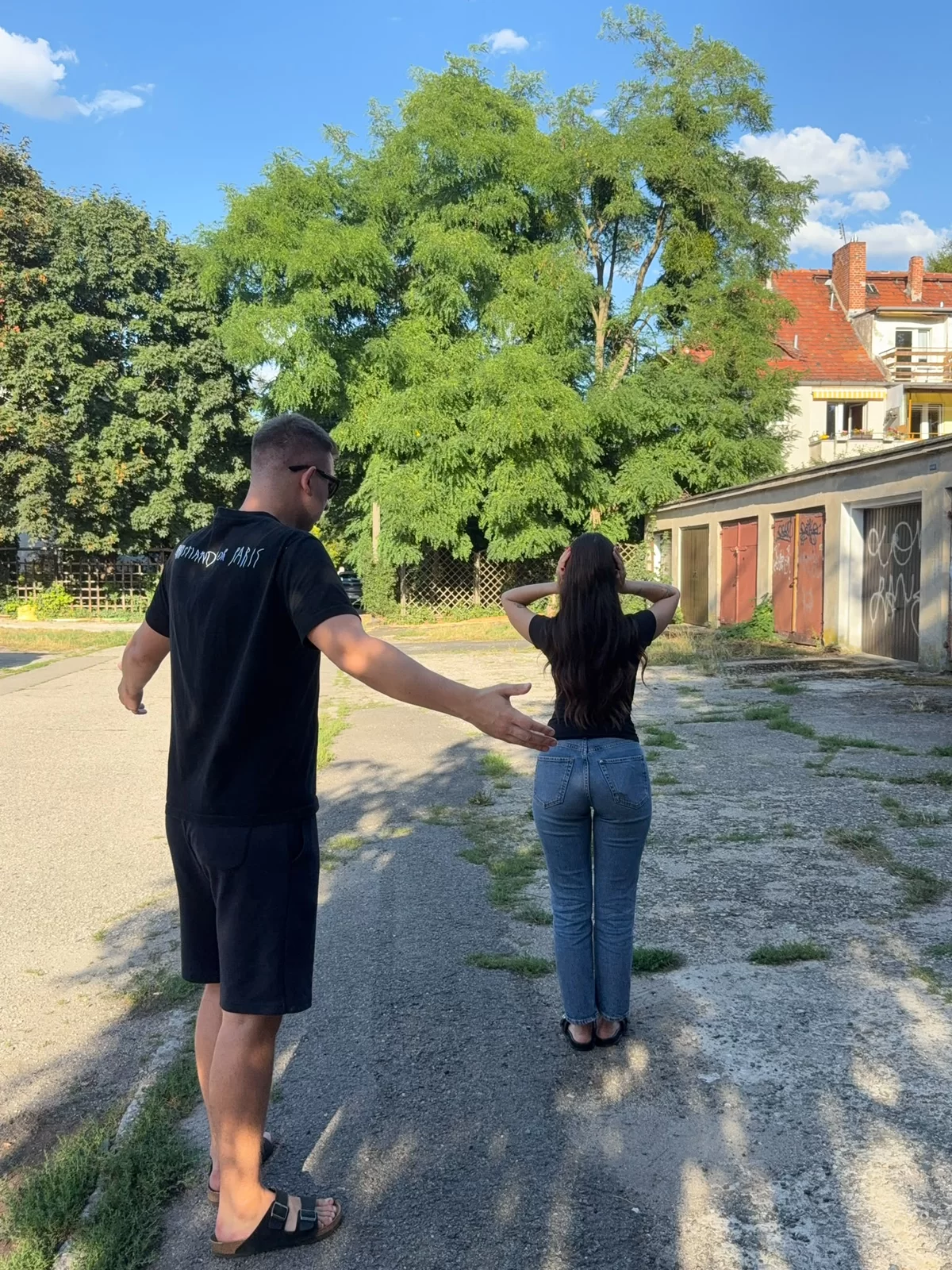 Słoneczny dzień, podwórko przy garażach. Na pierwszym planie stoi chłopak ubrany w czarną koszulkę, krótkie spodenki i klapki - ma rozłożone ręce. Przed nim stoi dziewczyna, ubrana w czarną koszulkę i niebieskie jeansy - dłonie przykłada do uszu - tak, żeby je zatkać.