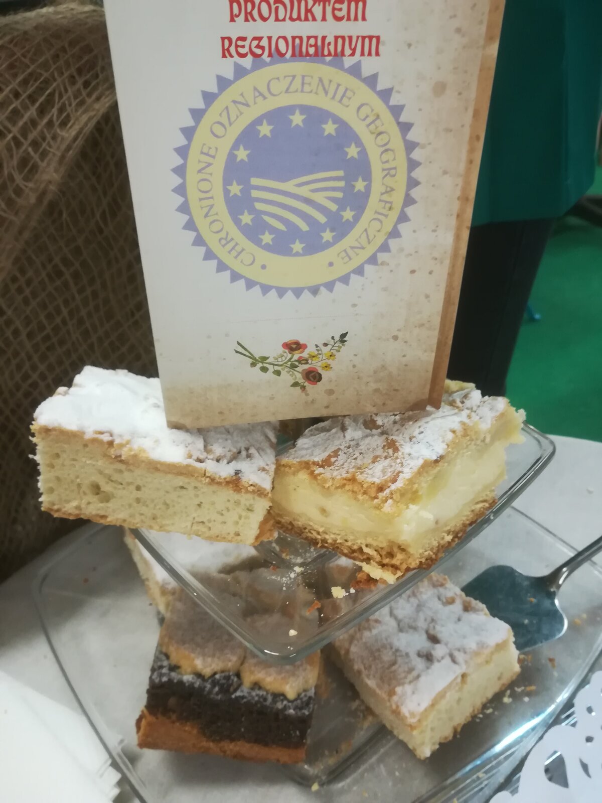 Na zdjęciu znajdują się kołacze śląskie. Kołacze to ciasto drożdżowe z nadzieniem, serowym, makowym lub jabłkowym. Na górze znajduje się tabliczka z informacją, że są to produkty regionalne ze znakiem chronionego oznaczenia geograficznego. 