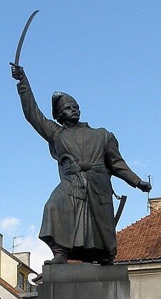 Fotografia przedstawia pomnik mężczyzny na postumencie, stojący w centrum miasta. Jest ubrany w długie szaty związane w pasie. Trzyma uniesioną do góry szablę, przy boku ma pistolet. W tle widać budynki mieszkalne.