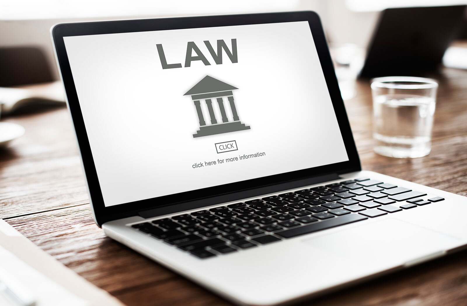  Zdjęcie przedstawia otwarty laptop stojący na drewnianym blacie. Na monitorze laptopa widoczna jest grafika przedstawiająca fasadę budynku z czterema kolumnami i spadzistym dachem. Powyżej widoczny jest napis w języku angielskim:LAW. Poniżej znajduje się okienko z napisem w języku angielskim. Nad nim widnieje napis:LAW, a pod budynkiem znajduje się przycisk:CLICK, oraz napis w języku angielskim:CLICK HERE FOR MORE INFORMATION. Na prawo od laptopa widoczna jest szklanka z wodą stojąca na blacie.