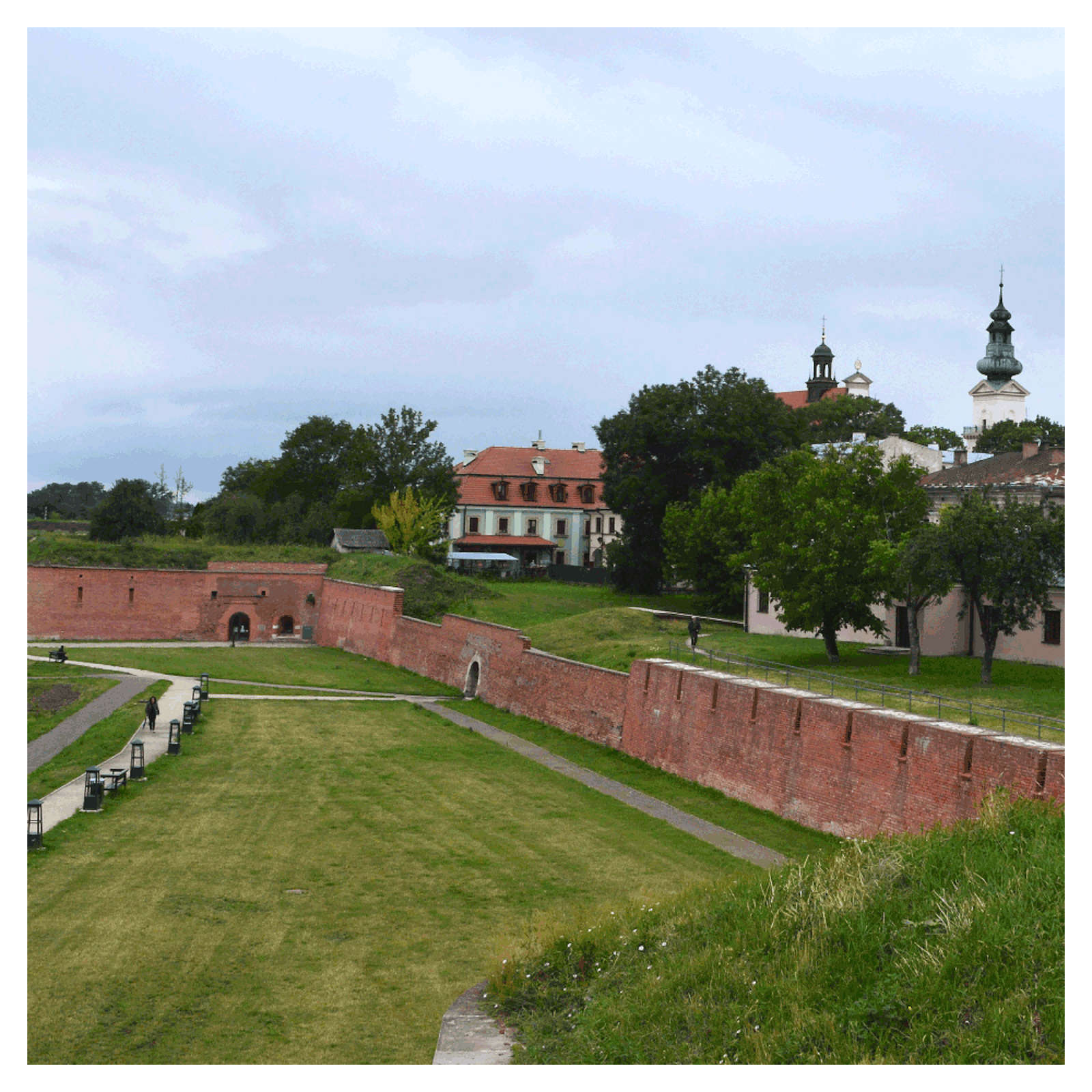 Zdjęcie przedstawia fragmenty fortyfikacji w Zamościu. To mury z cegły. U podnóża murów oraz powyżej nich rośnie trawa. Po prawej stronie zdjęcia zabytkowe budynki, w tym wieża kościoła. 