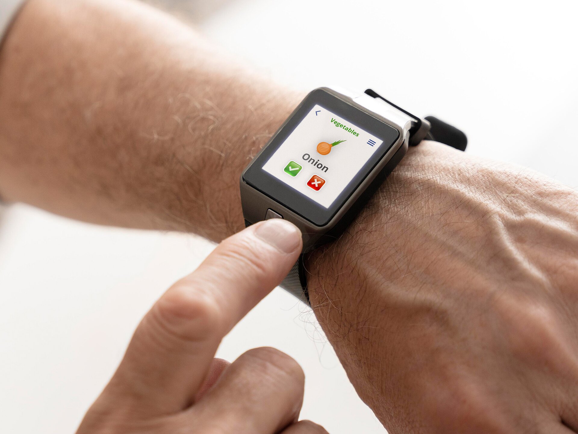Ilustracja przedstawia lewą rękę. Na nadgarstku widoczny jest duży zegarek z wyświetlaczem. Na wyświetlaczu widoczny jest napis w języku angielskim: Vegetables, poniżej którego widoczny jest rysunek cebuli. Poniżej znajduje się napis w języku angielskim: Onion oraz dwie ikony – po lewej zielony kwadracik z ptaszkiem, a po prawej czerwony kwadracik z iksem. Palec wskazujący prawej ręki dotyka przycisku na zegarku, który znajduje się na górnej części bransoletki zegarka.