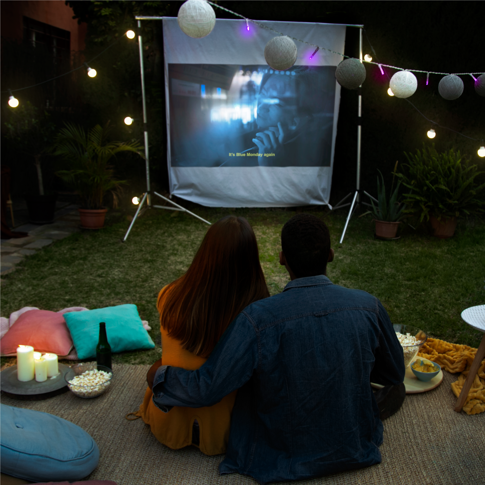 Zdjęcie przedstawia kobietę i mężczyznę siedzących na kocu w ogrodzie i oglądających film wyświetlany na rozwieszonej, białej płachcie.