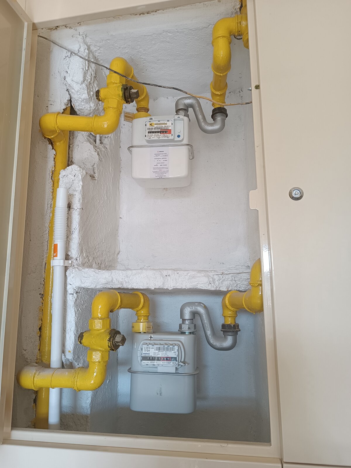 Na zdjęciu widoczne są dwa gazomierze miechowe zamontowane jeden pod drugim w gospodarstwie domowym. Gazomierz miechowy to urządzenie używane do pomiaru zużycia gazu w gospodarstwach domowych oraz innych budynkach. Przypomina niewielką metalową skrzynkę z wystającym okienkiem na przedniej stronie. Na okienku znajduje się kod kreskowy, informacje na temat urządzenia oraz licznik gazu. Do górnej części obu gazomierzy przymocowane są rury, którymi prowadzone jest paliwo gazowe.