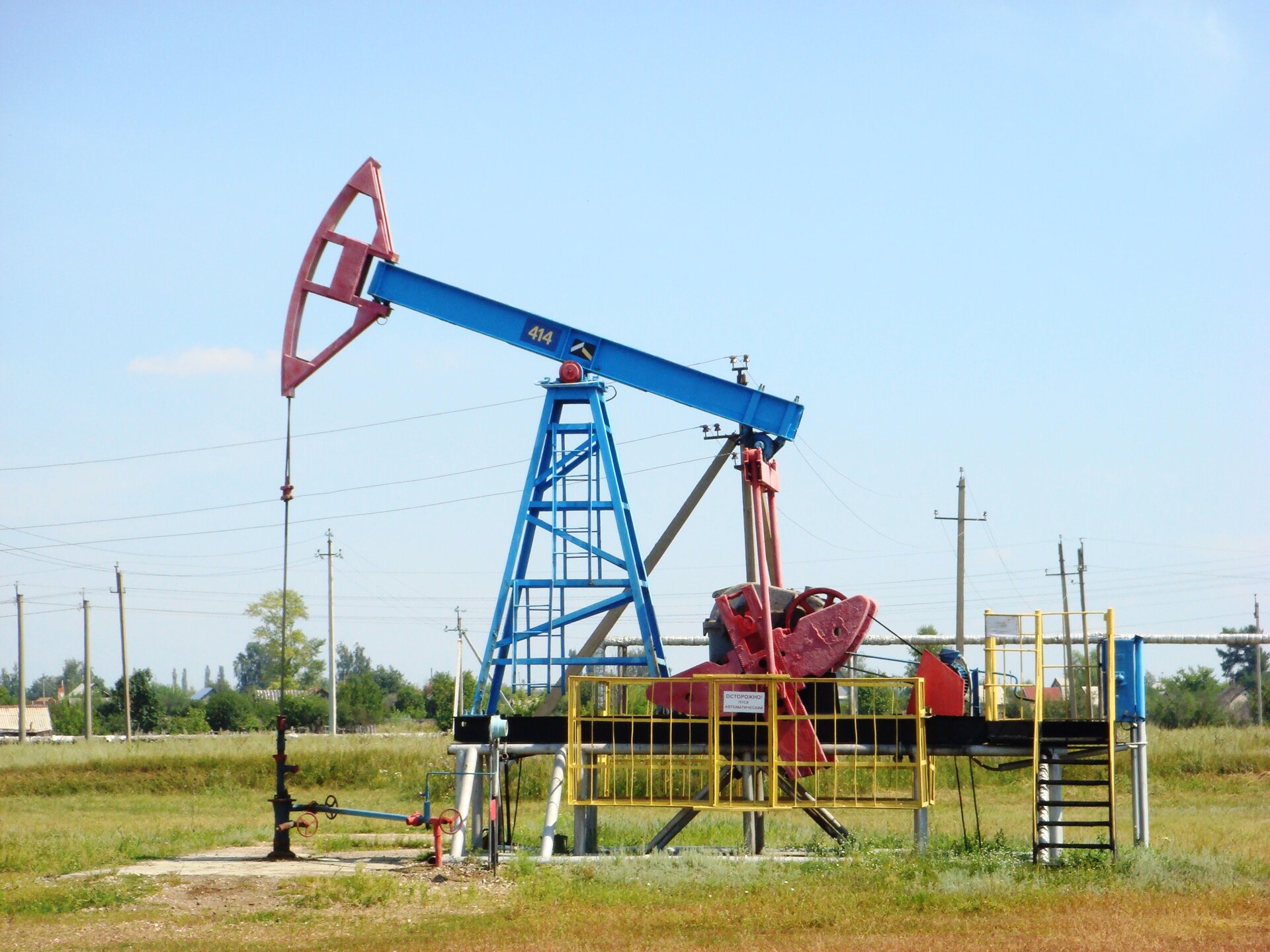 Na zdjęciu znajduje się urządzenie pompowe typu konwencjonalnego typ 7 SK 7. Kiwon jest rodzajem pompy wykorzystywanej do pompowania ropy naftowej ze złoża niskiego ciśnienia, montowana przy wylocie otworu wiertniczego z którego wydobywana jest ropa za pomocą pompy wgłębnej.W centralnej części zdjęcia umieszczona jest czarna prostokątna platforma wsparta na szarej ramie. Z prawej strony platformy znajdują się czarne schodki z żółtymi poręczami oraz niebieska skrzynka. Na środku platformy  znajdują się żółte poręcze z kratą. Za nimi widać szary silnik urządzenia wraz z czerwonymi przeciwwagami. Silnik jest o kształcie cylindra.W lewej części platformy znajduje się niebieska rama nośna w kształcie ostrosłupa prawidłowego czworokątnego. Pomiędzy jego ramionami znajdują się drabinki tego samego koloru. Na szczycie ramy umieszone jest niebieski ramię zwane wahaczem.Z prawej strony wahacza przymocowane są dwa czerwone pociągacze połączone z przeciwwagami.Z lewej strony wahacza znajduje się jego czerwony łeb. Za pomocą żerdzi dławikowej czyli prostego czarnego kształtu, łeb połączony jest z czarną głowicą odwiertu. Na głowicy znajdują się dwa czerwone zawory.Obok czarnej głowicy znajduje się druga mniejsza głowica koloru czerwonego. Posiada ona jeden czerwony zawór. Obie głowice połączone są niebieską rurą, która ma jeden czerwony zawór. Urządzenie znajduje się na polu, całe podłoże porośnięte jest trawą.W tle widać pojedyncze domy, drzewa oraz wiele słupów i linii wysokiego napięcia. Niebo jest koloru niebieskiego, nie ma chmur.