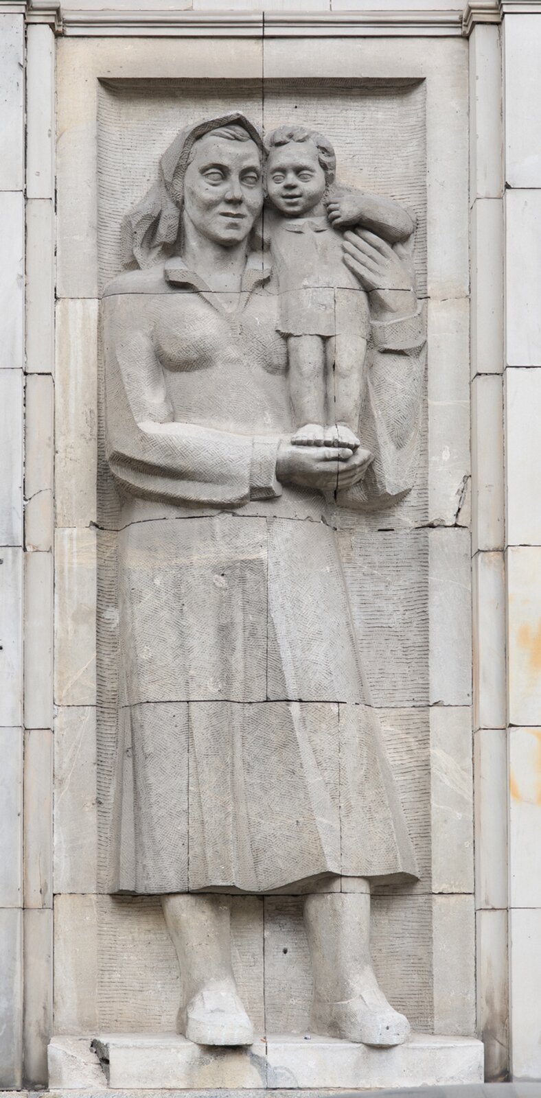 Fotografia przedstawia pładskorzeźbę, ukazującą matkę z dzieckiem na rękach. Kobeta ubrana jest w długa sukienkę i chustkę na głowie. Dziewczynka, w krótkiej sukience, skierowana jest twarzą do widza. Oboje są uśmiechnięci.