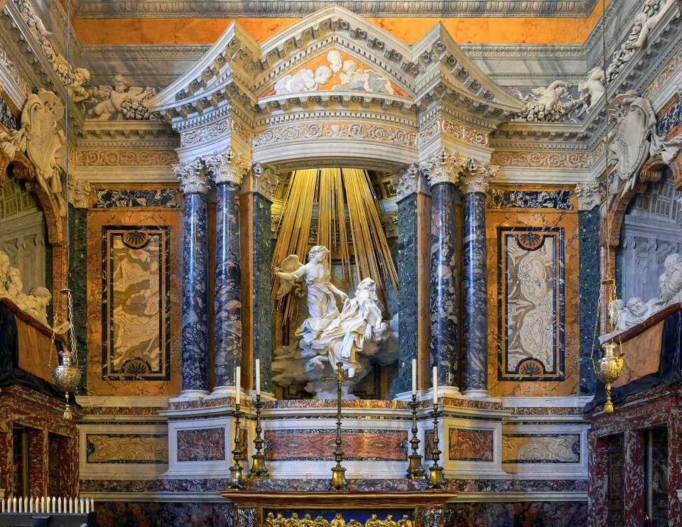 Zdjęcie przedstawia fragment barokowej kaplicy. W centrum w niszy mocno oświetlona grupa rzeźbiarska, ukazana na obłoku, z postaciami świętej Teresy i anioła. Kobieta ukazana jest w pozycji leżącej. Głowa świętej jest lekko przechylona w lewą stronę, do widza, ma ona półprzymknięte oczy i rozchylone usta, a jej lewa dłoń swobodnie opada wzdłuż ciała. Mocno udrapowana szata św. Teresy okrywa ją w całości, a jej całe ciało zwraca się w stronę strzały, którą trzyma w prawej dłoni stojący naprzeciw niej anioł i wymierza nią w postać świętej. Anioł stoi. Lewą dłonią dotyka jej szaty, przechylając lekko głowę w lewą stronę. Ma on kręcone, nieco dłuższe włosy, okryty jest lekko udrapowaną tkaniną, spod której widać prawy bark. Grupa rzeźbiarska wykonana została z białego marmuru. Na dwie postacie z góry spływają liczne złote promienie. Nisza, w której znajduje się rzeźba, wsparta jest na kolumnach o korynckich głowicach zdobionych liśćmi akantu. Po jej lewej i prawej stronie widoczne są podwójne kolumny również o korynckich głowicach i tak samo ozdobionych liśćmi akantu. Nisza ma kształt ośmiokąta i została wsparta na ołtarzu, a zwieńczona została bogato zdobioną attyką, nad którą widnieją białe stiuki z elementem dekoracyjnym. Pod nimi rzeźby amorków, a pomiędzy nimi elementy roślinne i owoce. Na ścianach bocznych liczne różnokolorowe marmury. Przed grupą rzeźbiarską znajduje się stół eucharystyczny, na którym ustawione są po lewej i po prawej stronie świeczniki, a na środku krzyż. Na wysokości centralnej grupy rzeźbiarskiej po lewej i prawej stronie w lożach, również wykonanych z marmuru, zostały umieszczone rzeźby ukazanych półpostaciowo członków rodziny Cornaro. Po prawej stronie od centrum zostało ukazanych siedemnaście niewielkich białych świeczek ustawionych w szeregu. 