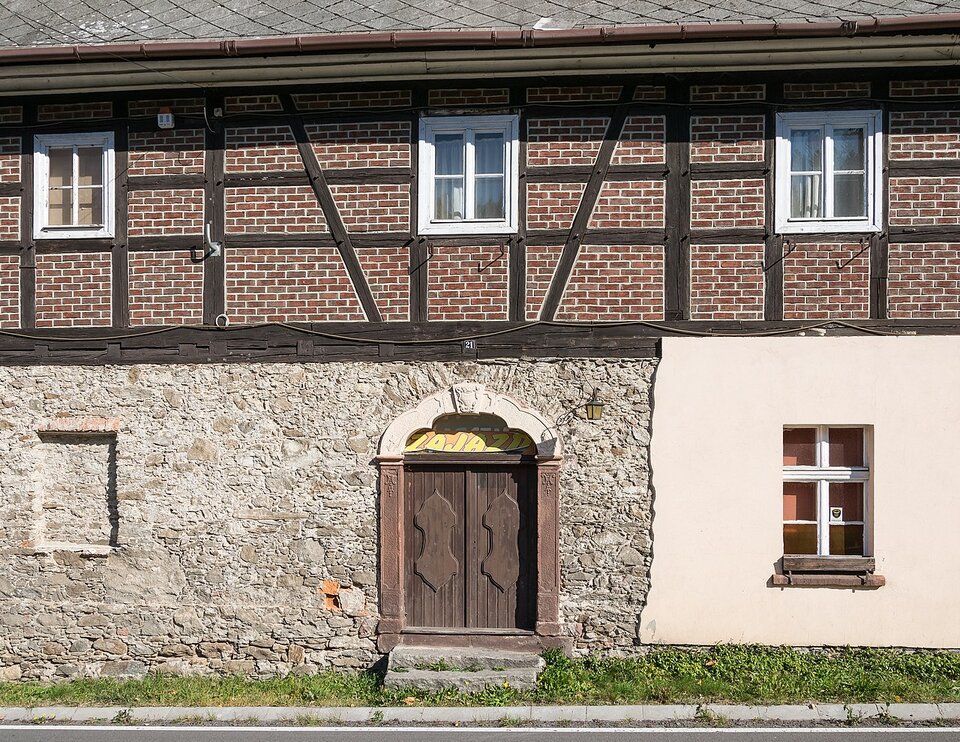 Zdjęcie ukazuje budynek. Budynek ma parter i piętro, niewielkie okna, spadzisty dach. Piętro wykonane jest z cegły. Między cegłami, w pewnych odstępach, są ułożone pionowo i poziomo drewniane belki. Po dwóch stronach jednego z okien, symetrycznie, belki ułożone są nieco na skos. Okno to znajduje się nad drzwiami budynku. Na parterze budynku część elewacji odpadła, odsłaniając ścianę wykonaną z kamienia.   