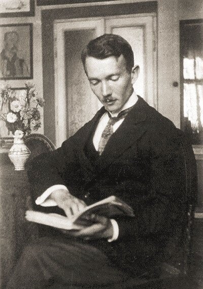 Zdjęcie przedstawia dojrzałego mężczyznę. Ma krótkie włosy oraz małe wąsy. Ubrany jest w surdut. Mężczyzna siedzi na krześle i czyta książkę. W tle okno, stolik z wazonem i kwiatami.