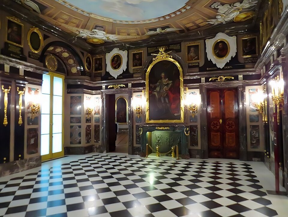 Zdjęcie przedstawia współczesny wygląd Pokoju Marmurowego na Zamku Królewskim w Warszawie z marmurowym kominkiem ozdobionym złoconymi dekoracjami. Na ścianie i pod sufitem widzą obrazy. Ściany są zdobione. Widać drzwi i okno. Podłoga jest w kratę.   