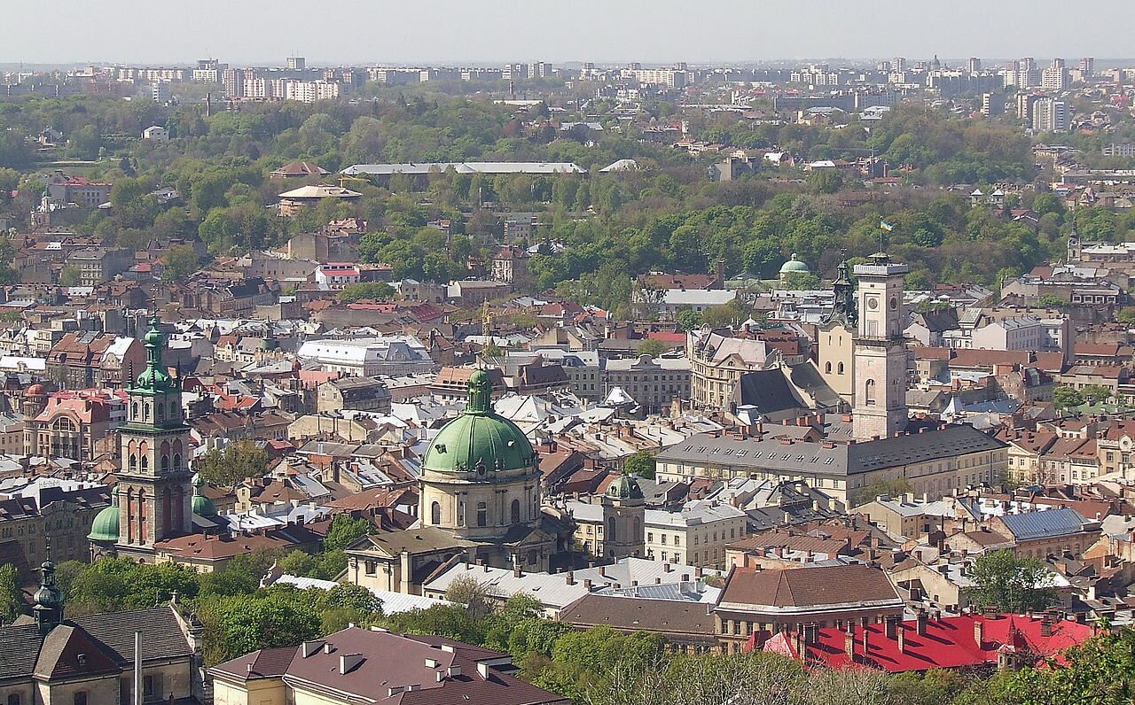 Zdjęcie przedstawia panoramę Lwowa. Zrobione jest z lotu ptaka. Znajdują się tu niezliczone budynki, kościoły, zabytki. Między nimi jest dużo zielonych drzew. Zabudowania sięgają aż po horyzont.