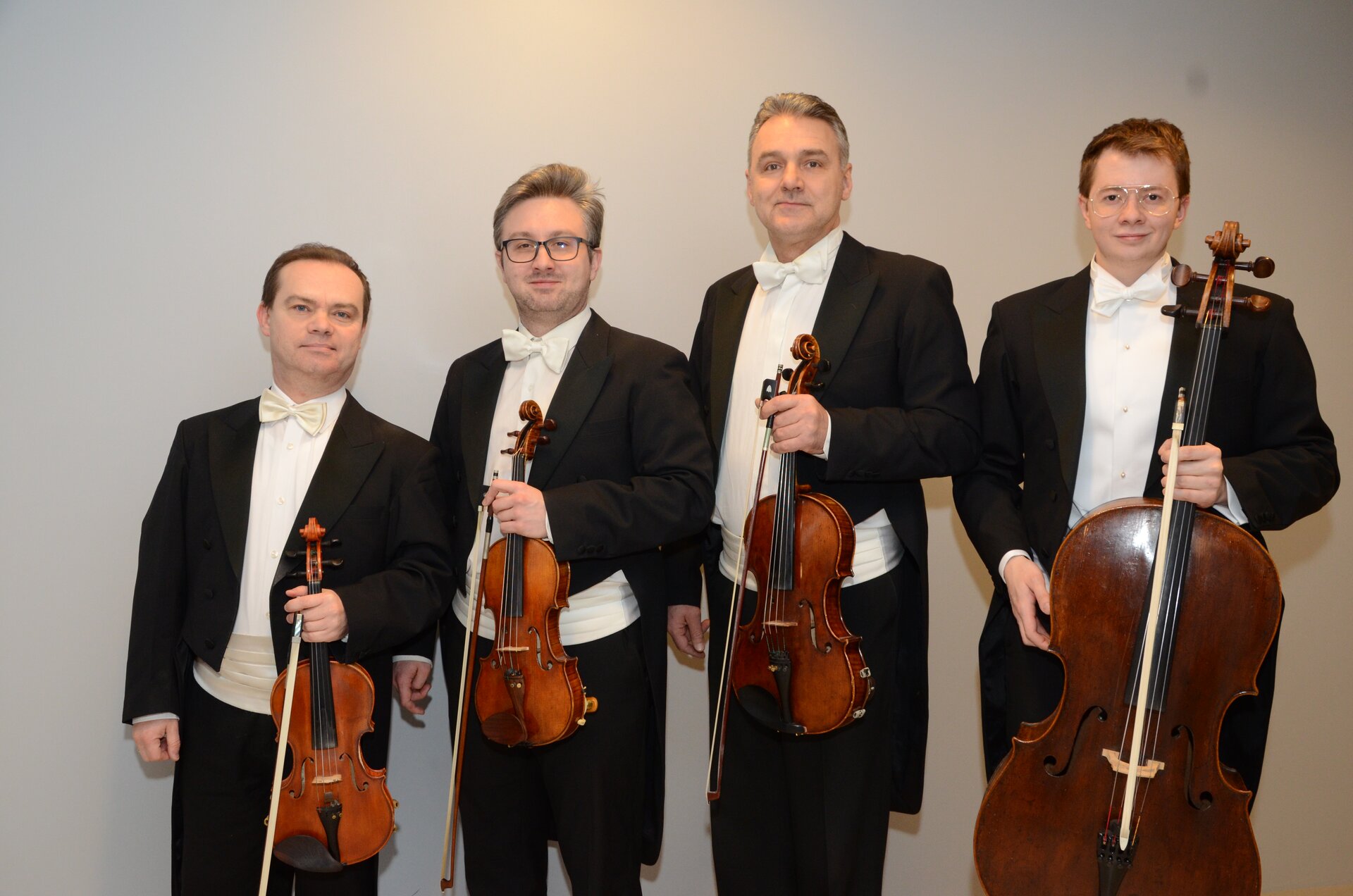 Fotografia przedstawia czterech muzyków ubranych w czarne fraki i białe koszule z białymi muchami. Mężczyźni stoją obok siebie na tle szarej ściany. W dłoniach trzymają instrumenty oraz smyczki, kolejno: skrzypce, skrzypce, altówkę oraz wiolonczelę. Mężczyźni mają uśmiechnięte twarze. 