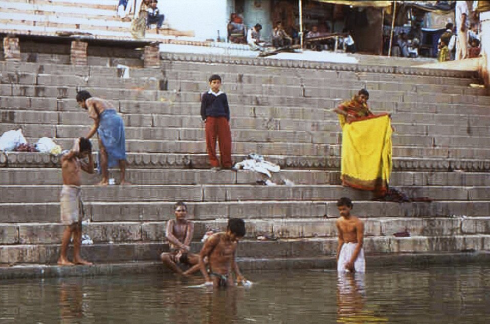 Zdjęcie przedstawia hinduistów kąpiących się w rzece Ganges. Przy brzegu kilkoro młodych chłopców umywa się i modli w świętej rzece. Niektórzy obserwują stojąc na schodach, inni składają ręczniki i ubrania.