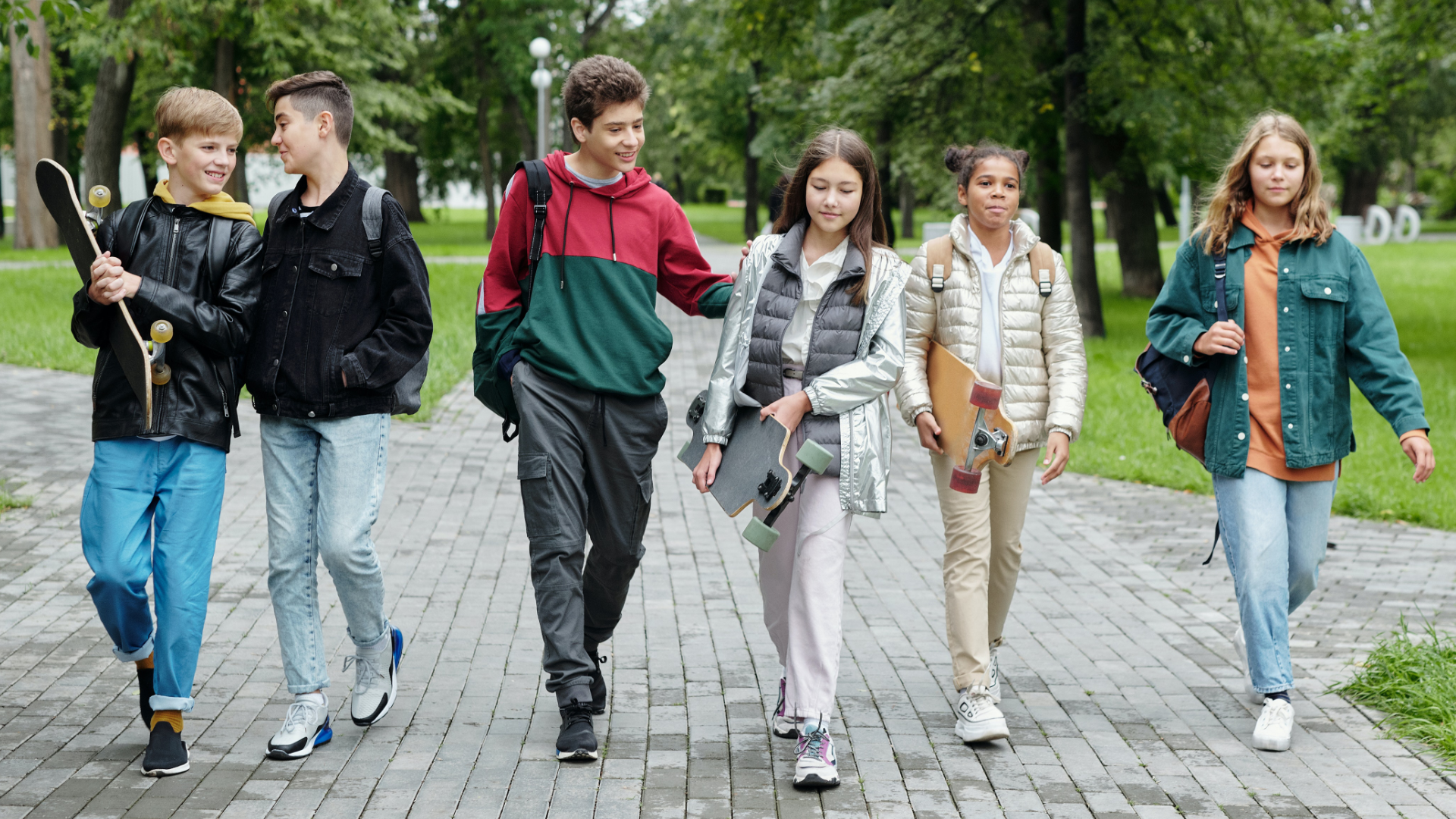 Zdjęcie przedstawia grupę sześciorga nastolatków, trzy dziewczyny i trzech chłopców. Idą chodnikiem, mają plecaki na plecach. Jedna z dziewczyn i jeden z chłopców niosą ze sobą deskorolki.