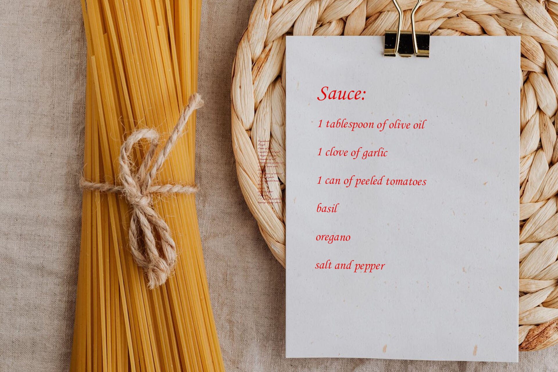 Zdjęcie przedstawia leżące na lnianej, szarej tkaninie: po lewej – makaron spaghetti związany szarym sznurkiem, a po prawej – kartkę przypiętą dużym spinaczem, leżącą na plecionej podkładce. Na kartce widoczny jest przepis na sos w języku angielskim: Sauce: 1 tablespoon of olive oil, 1 clove of garlic, 1 can of peeled tomatoes, basil, oregano, salt, and pepper.