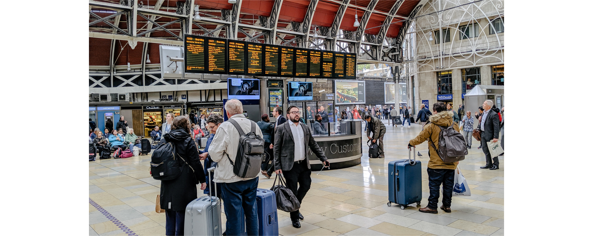 Zdjęcie przedstawia halę na lotnisku. Na zdjęciu widoczni są pasażerowie z bagażami, tablica przylotów i odlotów.