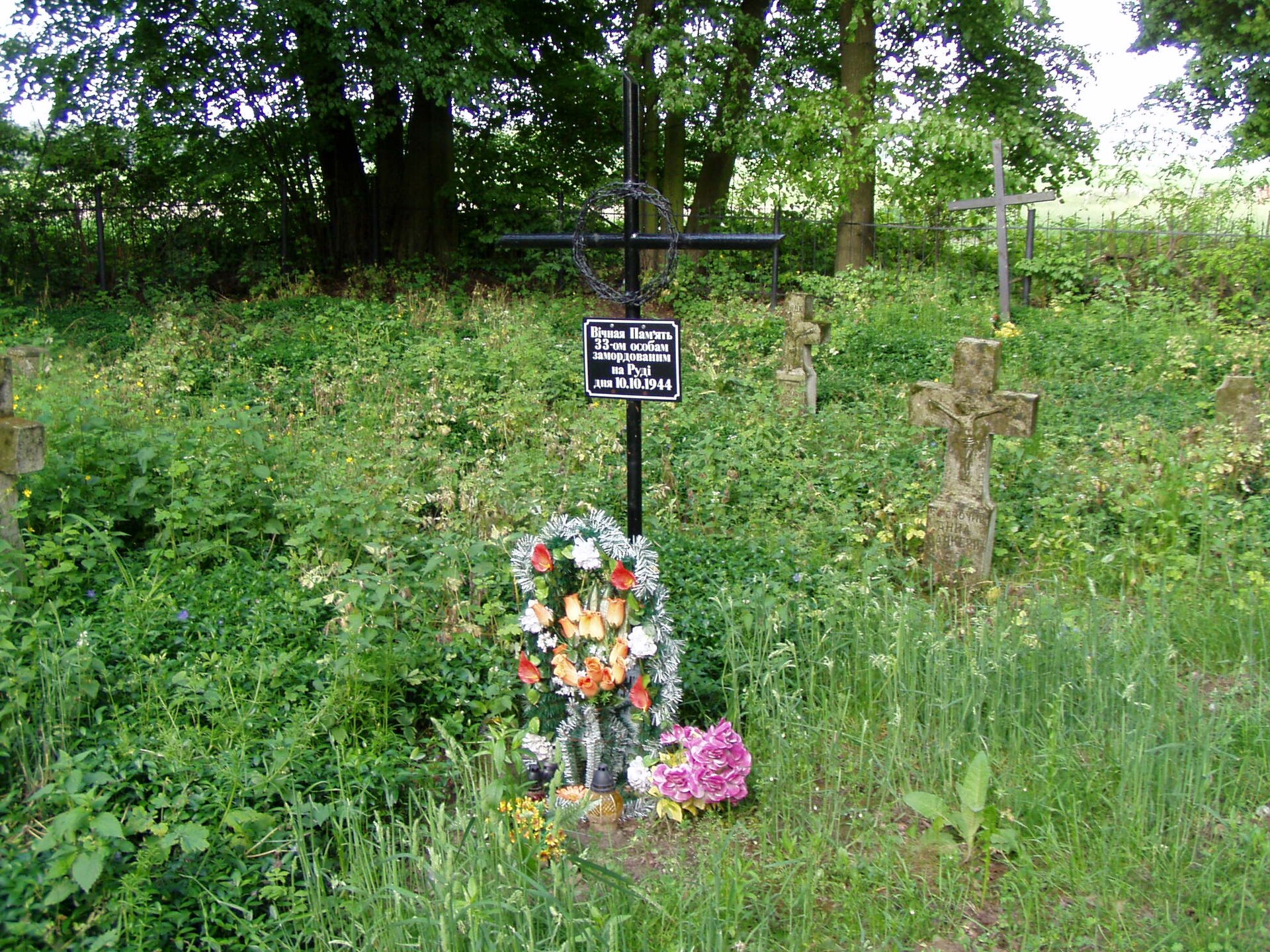 Zdjęcie przedstawia miejsce pamięci pomordowanych mieszkańców Starego Lublińca w otoczeniu innych mogił leśnych: kamiennych i drewnianych krzyży. Przy metalowym, czarnym krzyżu położone są wiązanki kwiatów. Na krzyżu wisi tabliczka pamiątkowa.