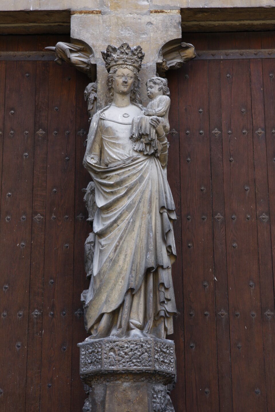 Ilustracja przedstawia fragment środkowego portalu zachodniego katedry w Reims. Ukazuje figurę Marii z Dzieciątkiem z filara międzyościeżowego. Maria ma na głowie koronę. Trzyma Jezusa na lewym przedramieniu. Jej szaty układają się swobodnie, przy czym jeden ich koniec trzyma w lewej dłoni. 