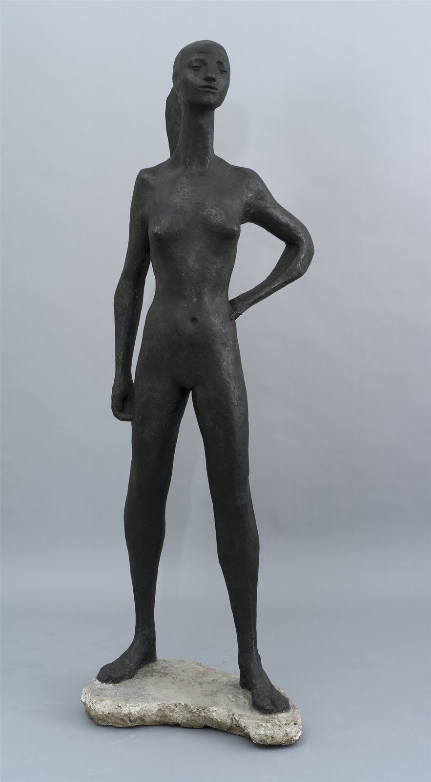 Fotografia przedstawia ciemnoszarą rzeźbę o jednolitej, chropowatej strukturze, uchwyconą na tle jasnej ściany. Rzeźba stoi na podstawie przypominającej płaską skałę o nieregularnych krawędziach, przedstawiając kobietę stojącą w rozkroku, trzymającą się lewą ręką pod bok. Prawa ręka nagiej postaci zwisa wzdłuż ciała. Kobieta jest szczupła, ma długą szyję oraz wyciąga do przodu głowę. Jej usta są wąskie, a oczy przymrużone. Z tyłu widać spięte włosy opadające na plecy.