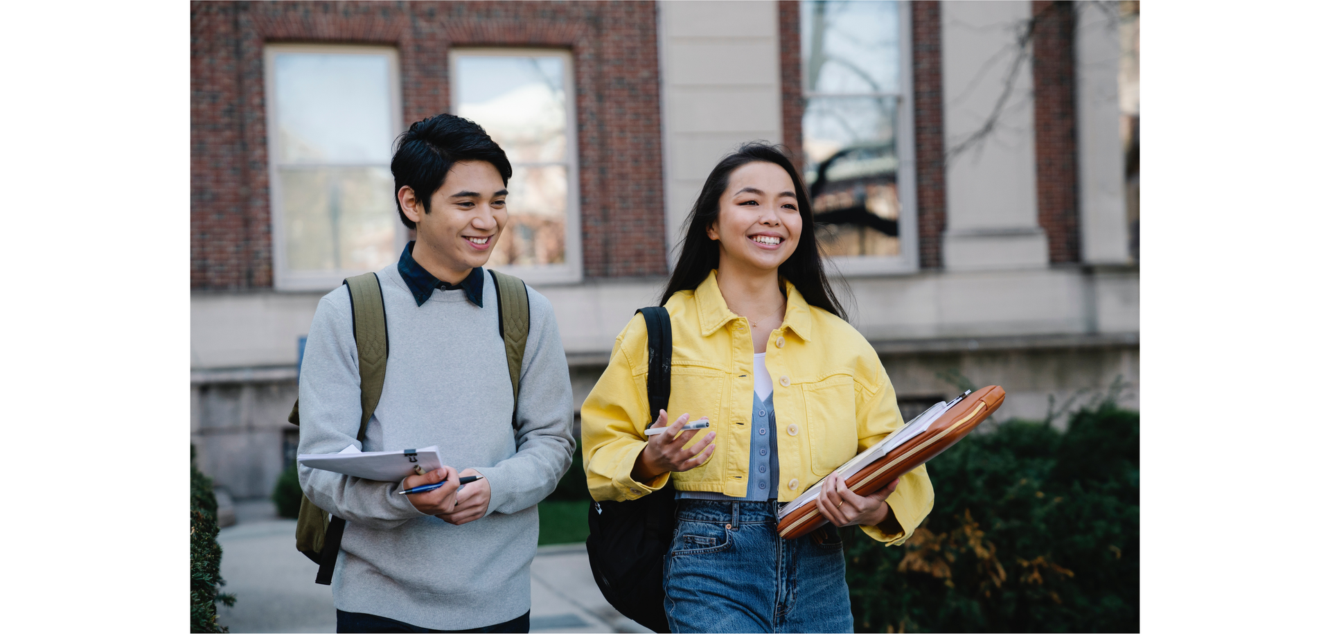 Zdjęcie przedstawia uśmiechniętych dziewczynę i chłopaka. W rękach niosą  jakieś notesy, na plecach mają plecaki.