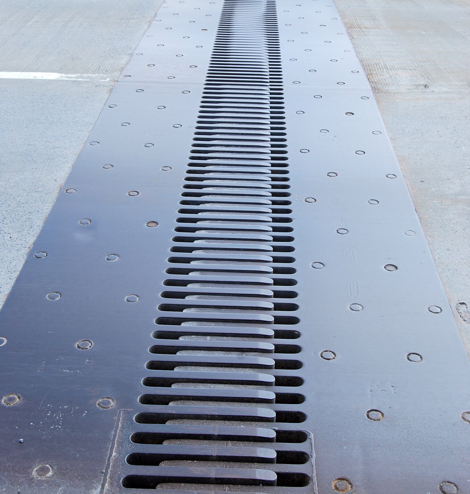 Rys. 3. Na zdjęciu przedstawiono fragment mostu. Most jest zbudowany z betonowych płyt, pomiędzy którymi pozostawiony jest pewien odstęp. Szerokość odstępu jest porównywalna z długością ludzkiej stopy. Na tym odstępie tym zamontowana jest szczelina dylatacyjna. Jest ona zbudowana z metalu pomalowanego na brązowo. Szczelina składa się z dwóch prostokątnych płyt zakończonych zaokrąglonymi zębami. Płyty szczeliny są zamocowane na betonowej płycie mostu za pomocą śrub, których główki są widoczne. Zęby obydwu szczelin wchodzą między siebie naprzemiennie, tak jak w zamku błyskawicznym. Zęby nie dotykają brzegu przeciwległej szczeliny – pozostaje tam trochę wolnego miejsca o długości zbliżonej do największego palca u stopy.